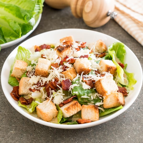 Caesar salad with crisp lettuce, croutons, bacon bits, and shredded cheese in a white bowl.