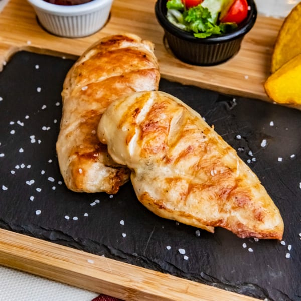 Two grilled chicken breasts with salt flakes on a black slate, with salad and sauce.