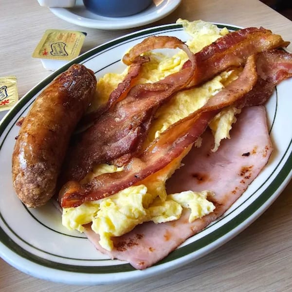 A diner breakfast plate with scrambled eggs, bacon, ham, and a sausage link.